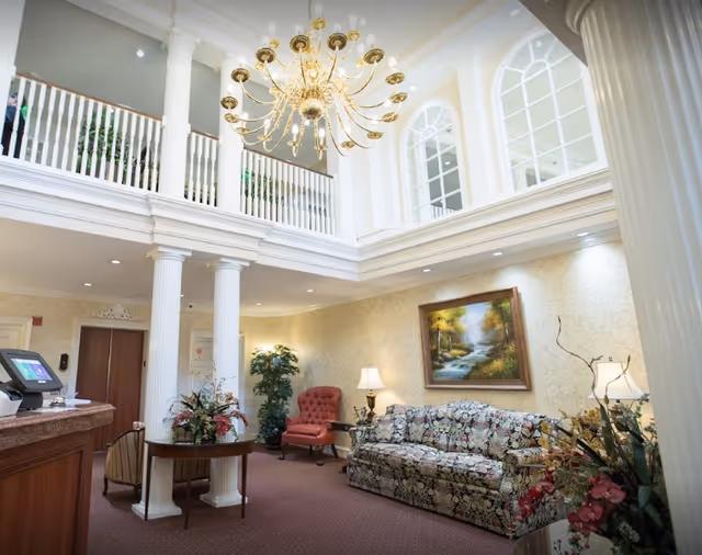 Two-story lobby lounge with tall white columns, a large chandelier, floral sofa, armchair, reception desk and a framed landscape painting.
