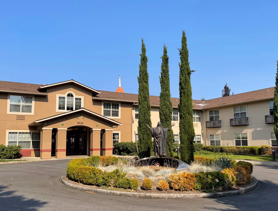 Exterior view of Assumption Village senior living facility showing a two-story building with beige and brown walls, multiple windows, and a covered entrance. In front of the building is a circular landscaped area with bushes, tall cypress trees, and a statue of a person with outstretched arms.