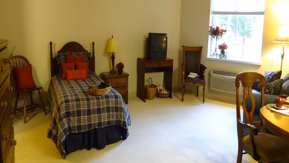 A cozy single bedroom with a twin bed, nightstand and lamp, TV on a small table, chairs and a dining table beside a window.