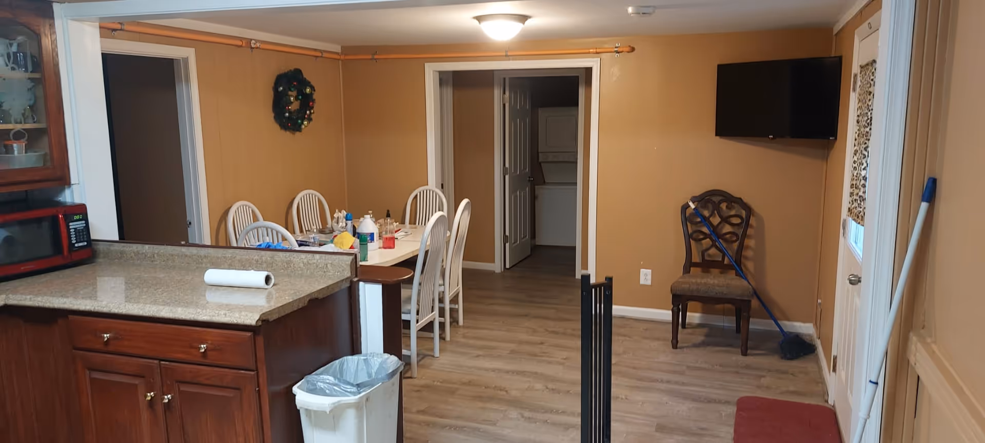 Interior view of a room in an assisted living facility showing a kitchen counter with a red microwave and paper towel roll, a dining table with six white chairs and various cleaning supplies on it, a doorway leading to a laundry area with a stacked washer and dryer, a wall-mounted TV, a wooden chair with a broom leaning against it, and cleaning tools near a door with a leopard print curtain.