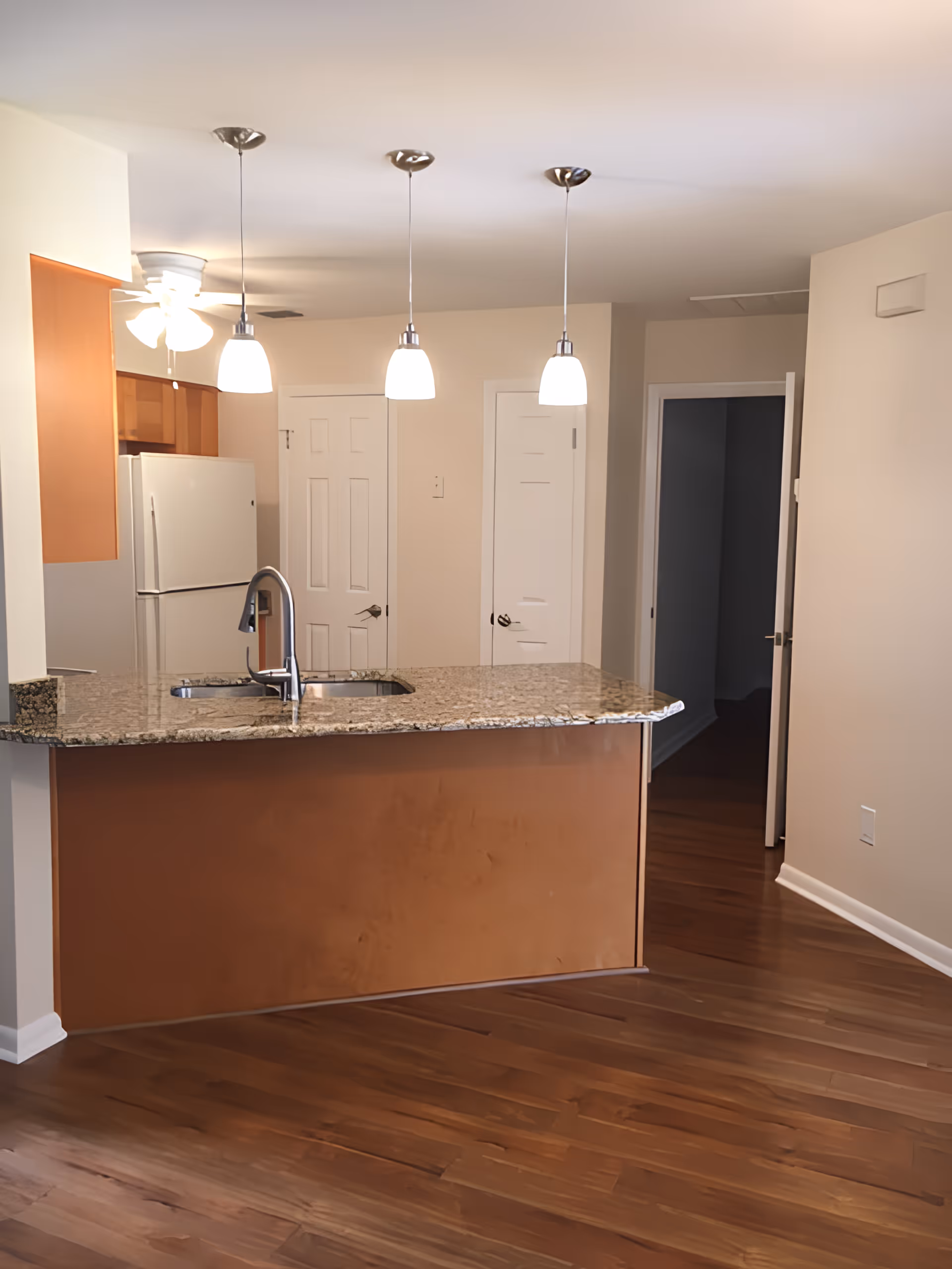 Interior view of a kitchen area featuring a granite countertop island with a built-in sink and faucet. Three pendant lights hang above the island. In the background, there is a white refrigerator, wooden cabinets, and two closed white doors. The floor is wooden, and the walls are painted beige.