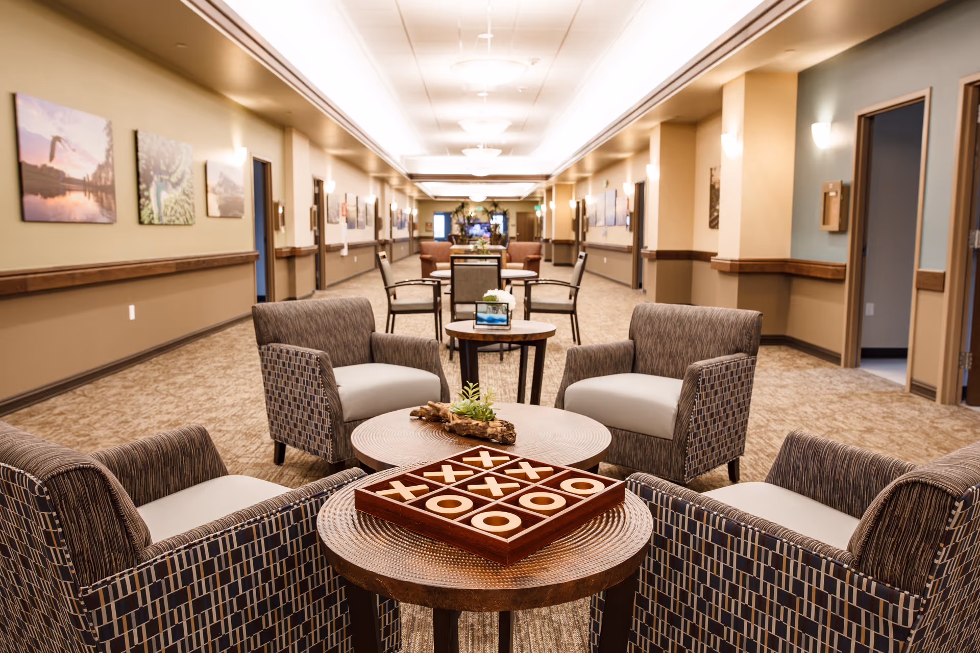 Well-lit memory care lounge with upholstered chairs and small tables arranged along a long hallway, a tic-tac-toe game visible on the nearest table.
