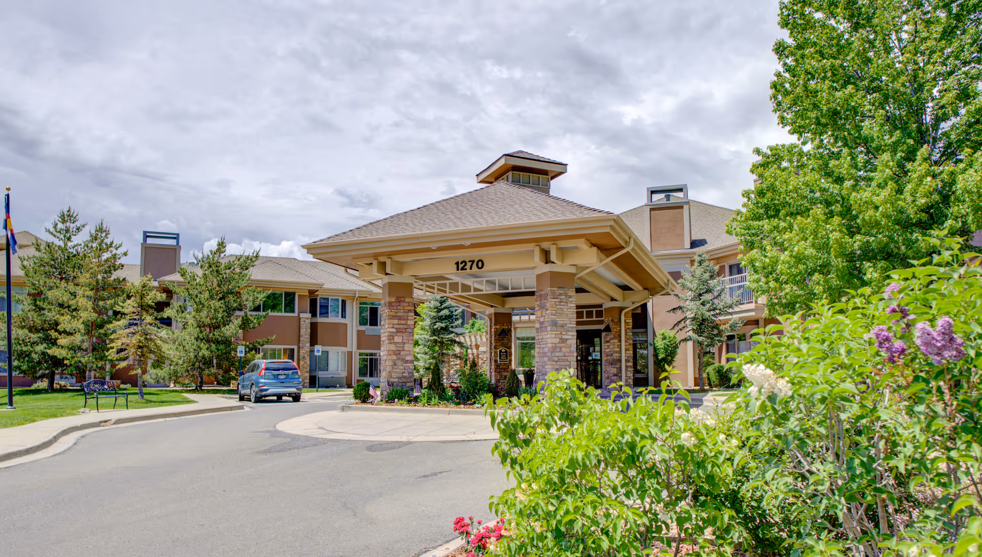 Entrance of a senior living facility with a covered drop-off area supported by stone pillars, surrounded by greenery and trees, with a driveway and a parked car nearby. The building has multiple windows and a beige exterior.
