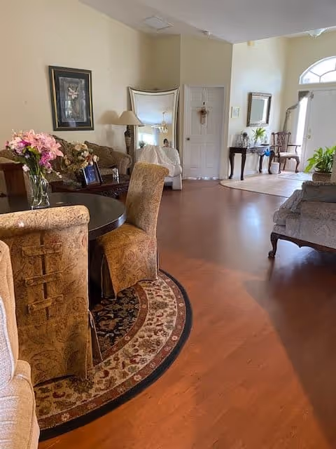A cozy living room area with wooden flooring, a round table with floral upholstered chairs, a vase of pink flowers on the table, a sofa, a large mirror leaning against the wall, a framed picture, a lamp, and a hallway leading to a door with a small wreath. There is also a console table with a mirror and plants near the entrance door.
