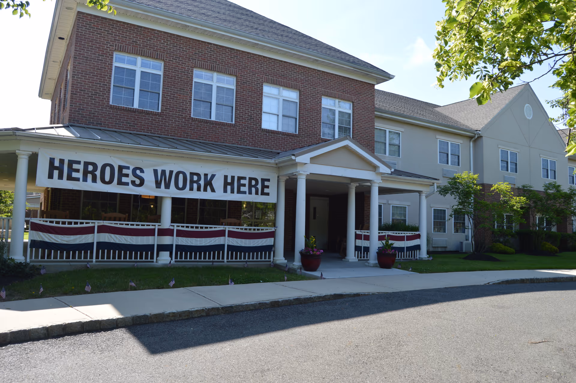 Exterior view of a senior living facility named Rose Hill with a brick and beige facade. A large banner reading 'HEROES WORK HERE' is displayed on the front porch area, which is decorated with red, white, and blue bunting. There are potted plants near the entrance and small American flags planted in the grass along the sidewalk.