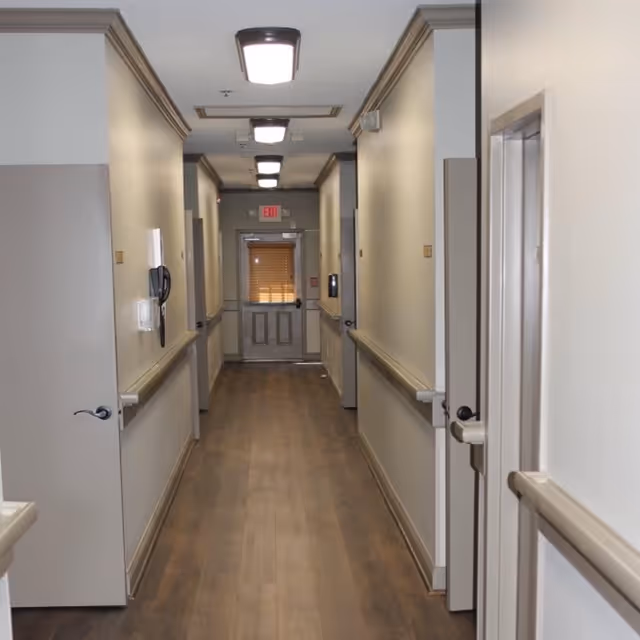 A clean, well-lit hallway in an assisted living facility with beige walls, wooden flooring, handrails on both sides, several closed doors, ceiling lights, and an exit door with a window at the end.