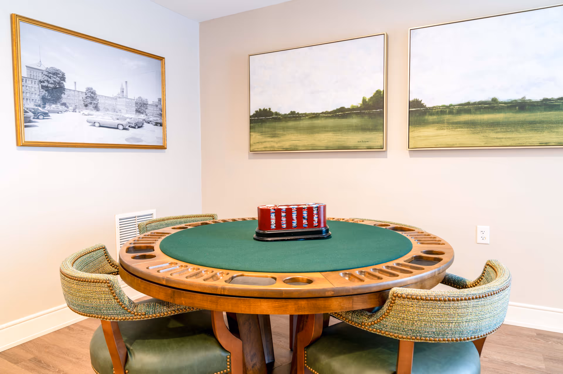 A round wooden game table with a green felt surface and built-in cup holders, surrounded by four upholstered chairs with green cushions. On the table is a red poker chip holder filled with poker chips. The walls have three framed pictures, one black and white photo of a building and two landscape paintings with green fields and cloudy skies.