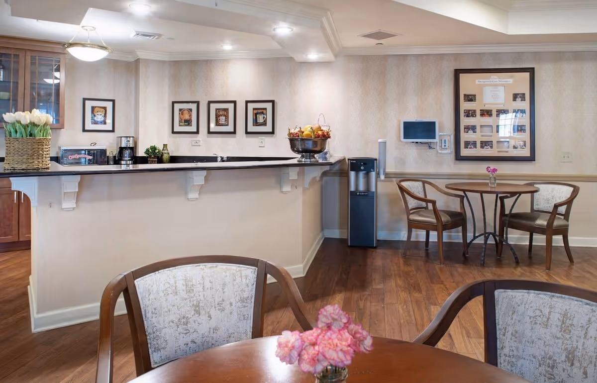 A cozy common area with wooden floors, a counter with a coffee maker, a fruit bowl, and framed pictures on the wall. There are two small round tables with chairs, each table decorated with a small vase of flowers. A water dispenser and a wall-mounted screen are also visible.
