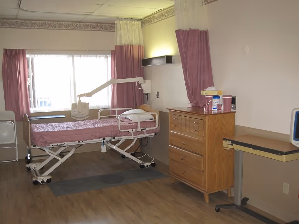 A patient room in a healthcare facility with a hospital bed covered in a pink blanket, a wooden dresser with various items on top, a small adjustable table on wheels, and a window with pink curtains letting in natural light.