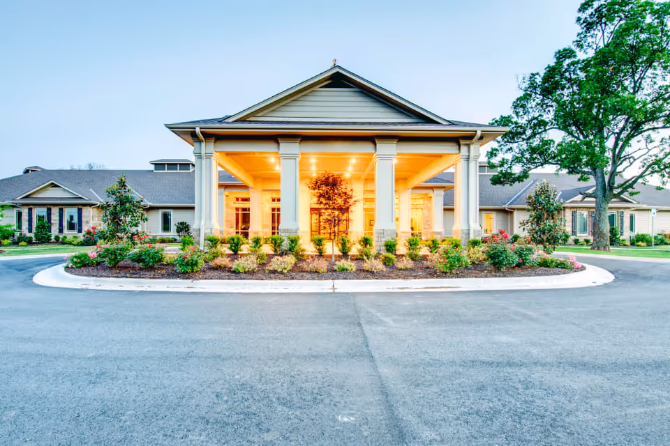 Front exterior view of Village on the Park Bentonville building with a covered entrance supported by columns, surrounded by landscaped bushes and trees, under a clear sky.