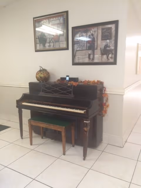 An upright piano with a wooden bench placed in front of it, decorated with a small globe and autumn-themed garland on top. Two framed pictures hang on the wall above the piano. The setting appears to be a clean, tiled indoor hallway or common area.
