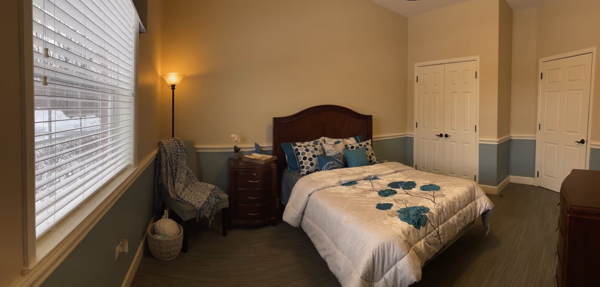 A cozy bedroom with a bed featuring a floral comforter and multiple pillows. There is a wooden nightstand with a lamp and some books next to the bed. A chair with a blanket draped over it sits near a window with blinds. The room has two closed white doors and a wooden dresser. The walls are painted beige with a blue wainscoting, and the floor is covered with wood-like flooring.