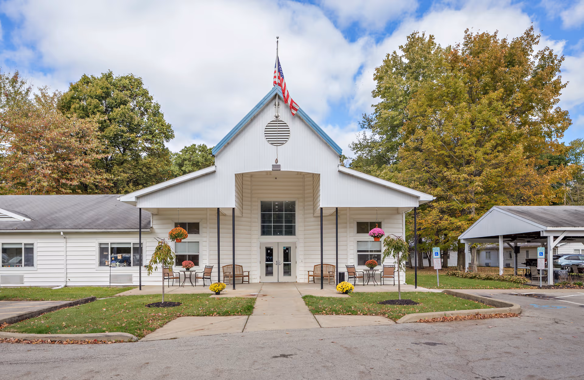 Front exterior view of The Grove at Greenville facility with a white building featuring a peaked roof and an American flag on top. There are benches and tables with chairs on the porch, potted flowers, and trees with autumn foliage surrounding the building. A covered parking area is visible to the right.