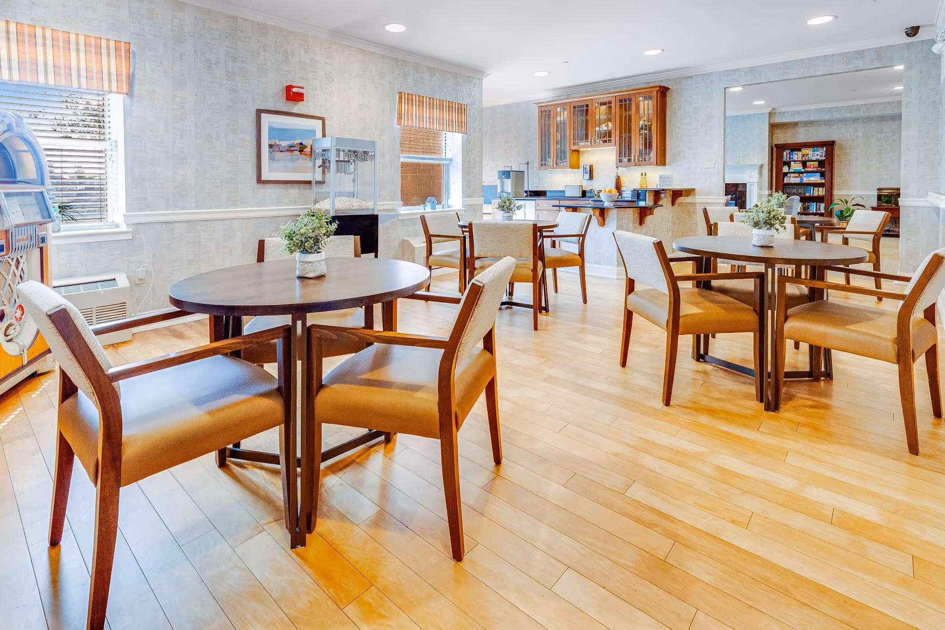 Bright communal dining area with round tables and chairs, wood floors, and a small kitchenette/bar in the background.