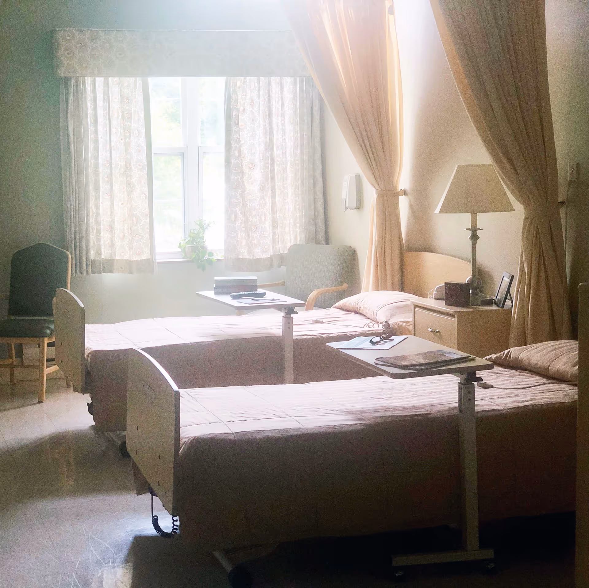 A softly lit room with two hospital-style beds, each with a beige bedspread and an adjustable overbed table. There is a window with patterned curtains letting in natural light, a green armchair, a wooden nightstand with a lamp, and beige privacy curtains partially drawn around one bed.