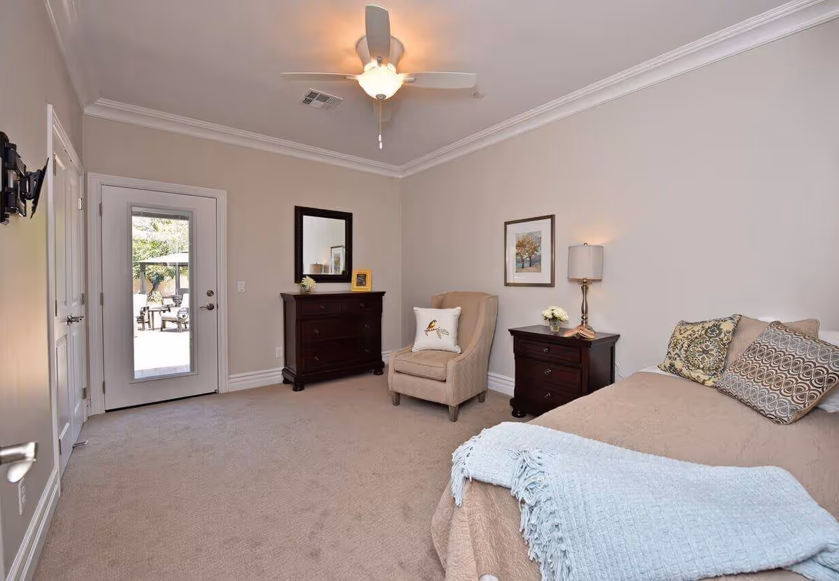 A cozy bedroom with beige walls and carpet. The room features a bed with multiple decorative pillows and a light blue throw blanket. Next to the bed is a dark wooden nightstand with a lamp and a small flower arrangement. There is a beige armchair with a bird-themed pillow, a dark wooden dresser with a mirror above it, and a door with a glass panel leading outside to a patio area with outdoor furniture. A ceiling fan with a light is mounted on the ceiling.
