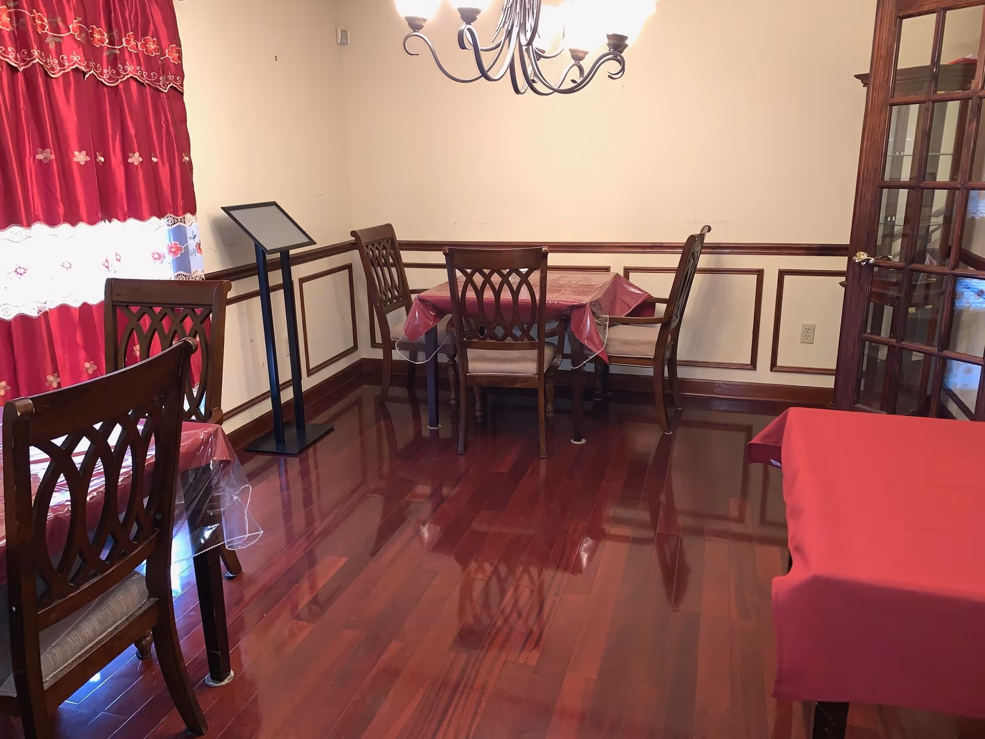 A dining room with polished wooden floors, a chandelier hanging from the ceiling, and several wooden chairs around tables covered with red and clear plastic tablecloths. There is a window with red curtains on the left and a glass-paneled door on the right. A black stand is positioned near the corner of the room.