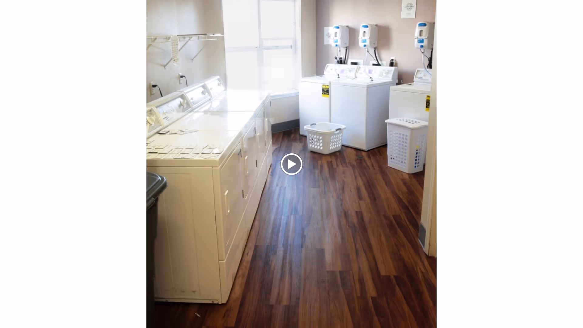 Laundry room with multiple white washing machines and dryers along the walls, two laundry baskets on the wooden floor, and a window letting in natural light.