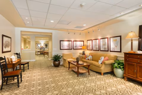 A well-lit interior common area with a beige patterned carpet, a tan sofa with colorful pillows, a wooden coffee table, an armchair, framed artwork on the walls, potted plants, and wooden chairs and furniture. The space has recessed lighting and a warm, inviting atmosphere.