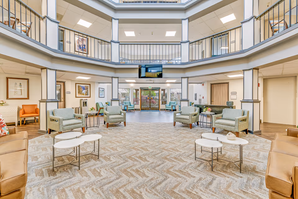 Bright and spacious senior living facility common area with modern furniture including green armchairs, brown leather sofas, and white nested coffee tables on a patterned carpet. The area features a two-story ceiling with a balcony railing above, framed artwork on the walls, and a large TV mounted on the wall. Glass doors lead outside, allowing natural light to fill the space.