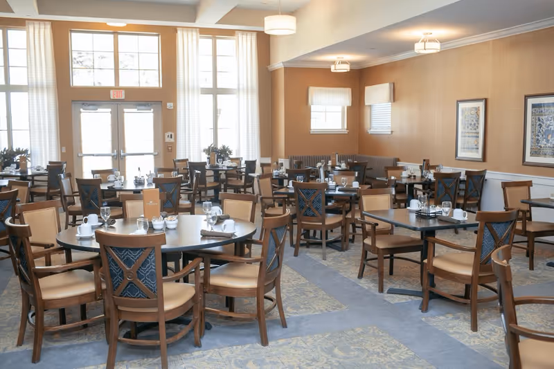 A spacious dining room with multiple round and square tables set with cups, glasses, and napkins. The room has large windows with white curtains, beige walls, framed artwork, and ceiling lights. Wooden chairs with cushioned seats surround the tables.