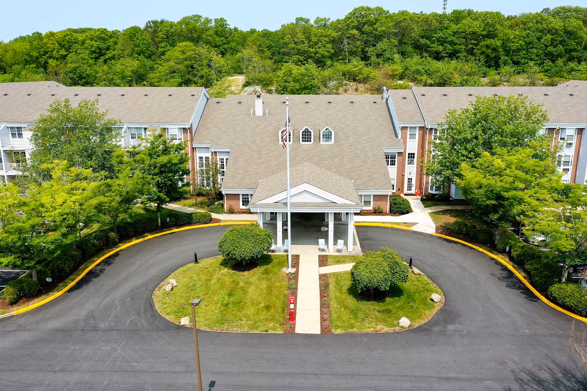 Aerial view of a senior living building's front entrance with a covered portico, circular driveway, flagpole, and landscaped greenery.