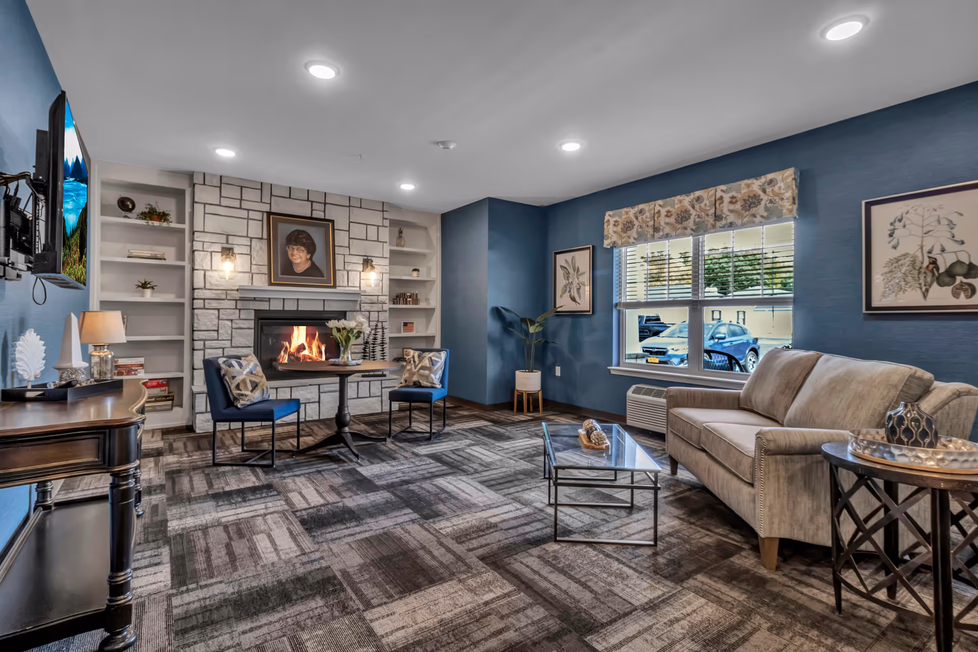 A cozy living room with blue walls, a patterned carpet, and a stone fireplace with a lit fire. Above the fireplace is a framed portrait. Two blue chairs with patterned pillows sit around a small round table with a vase of flowers. A beige sofa is positioned near a window with floral valance and blinds. There are framed botanical prints on the walls, a glass coffee table, and a wooden side table with decorative items. A flat-screen TV is mounted on the wall opposite the sofa.