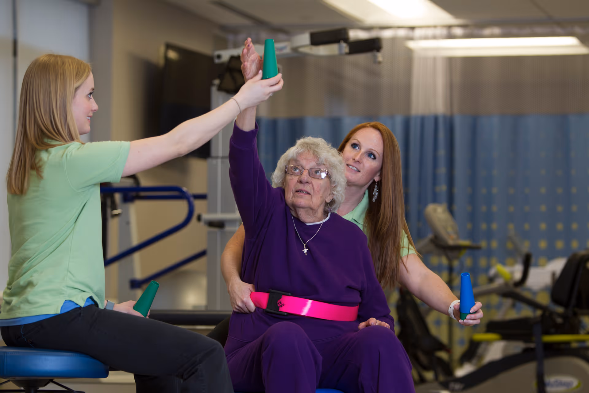An elderly woman in a purple outfit is seated and participating in a physical therapy exercise with the assistance of two female therapists. One therapist is helping her raise her arm while holding a green cone, and the other therapist is supporting her from behind, also holding a green cone. The setting appears to be a rehabilitation or therapy room with exercise equipment and privacy curtains in the background.
