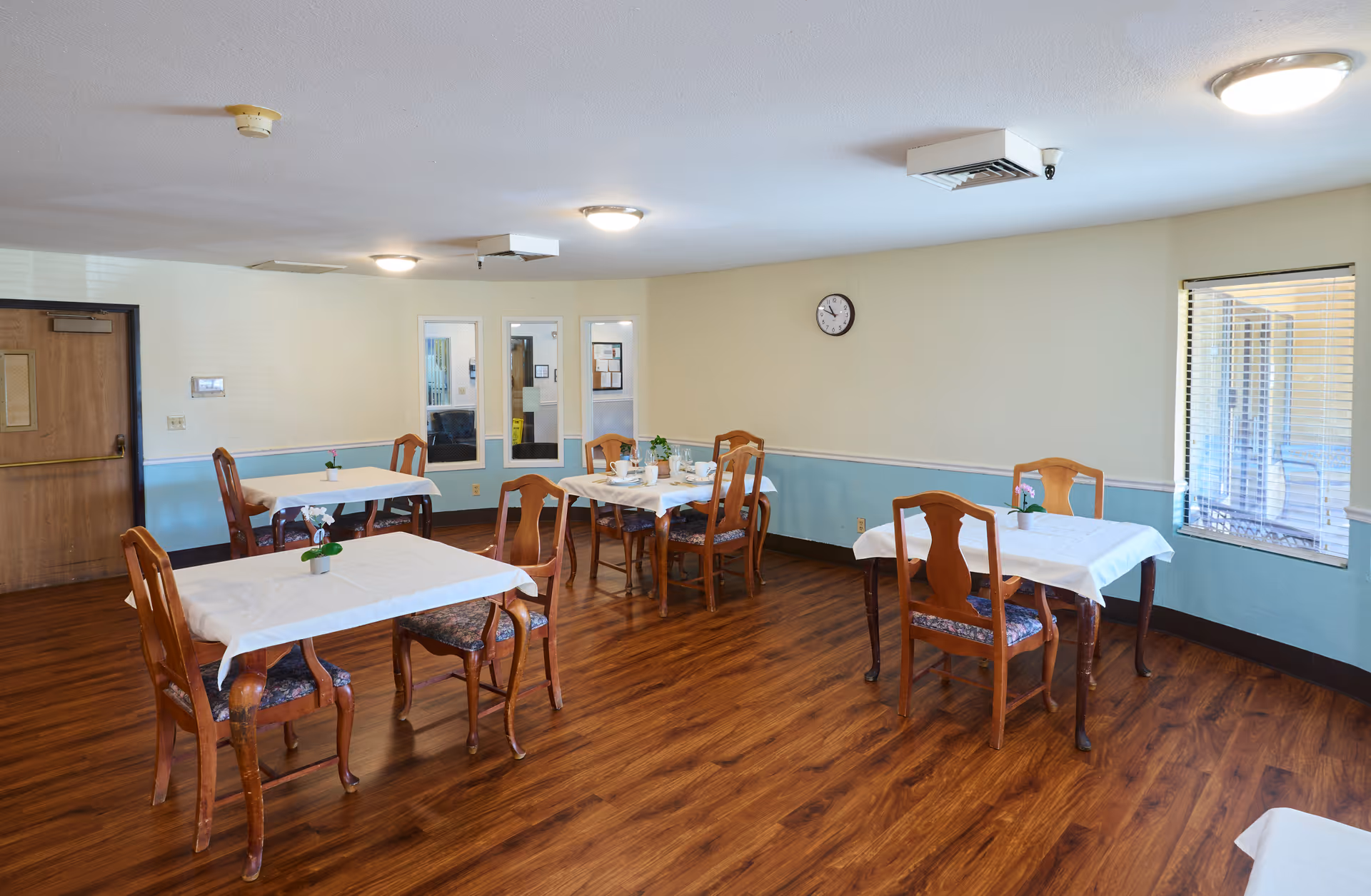 A dining room with wooden floors and light blue and cream walls. There are four tables covered with white tablecloths, each surrounded by wooden chairs with patterned cushions. Small potted plants are placed on each table. A clock is mounted on the wall, and windows with blinds allow natural light into the room.
