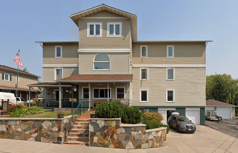 Three-story beige senior living building with entrance stairs and landscaped front yard.
