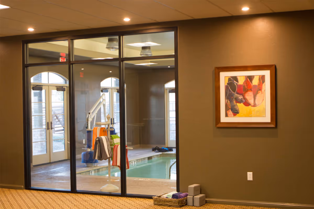 Indoor view of a senior living facility showing a glass wall separating a carpeted room with a framed colorful painting on the wall from an indoor pool area with pool lift and towels hanging on a stand.