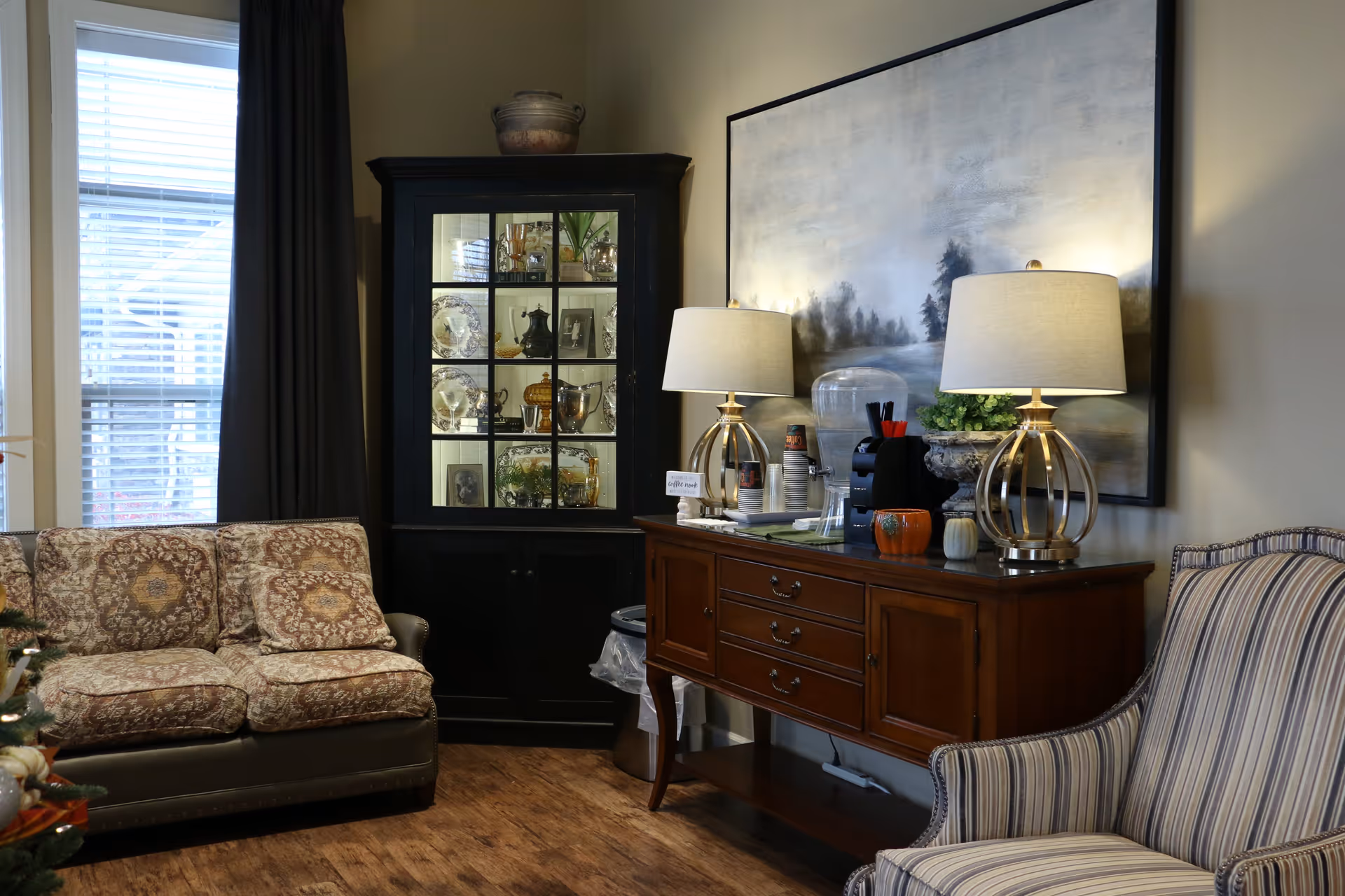 A cozy living room area with a patterned sofa on the left, a striped armchair on the right, and a wooden sideboard in the center. The sideboard holds two table lamps with beige shades, a water dispenser, a coffee machine, and small decorative pumpkins. Behind the sideboard is a large framed painting of a misty landscape. To the left of the sideboard is a black cabinet with glass doors displaying various decorative items and photos. A window with blinds and dark curtains is visible on the far left.