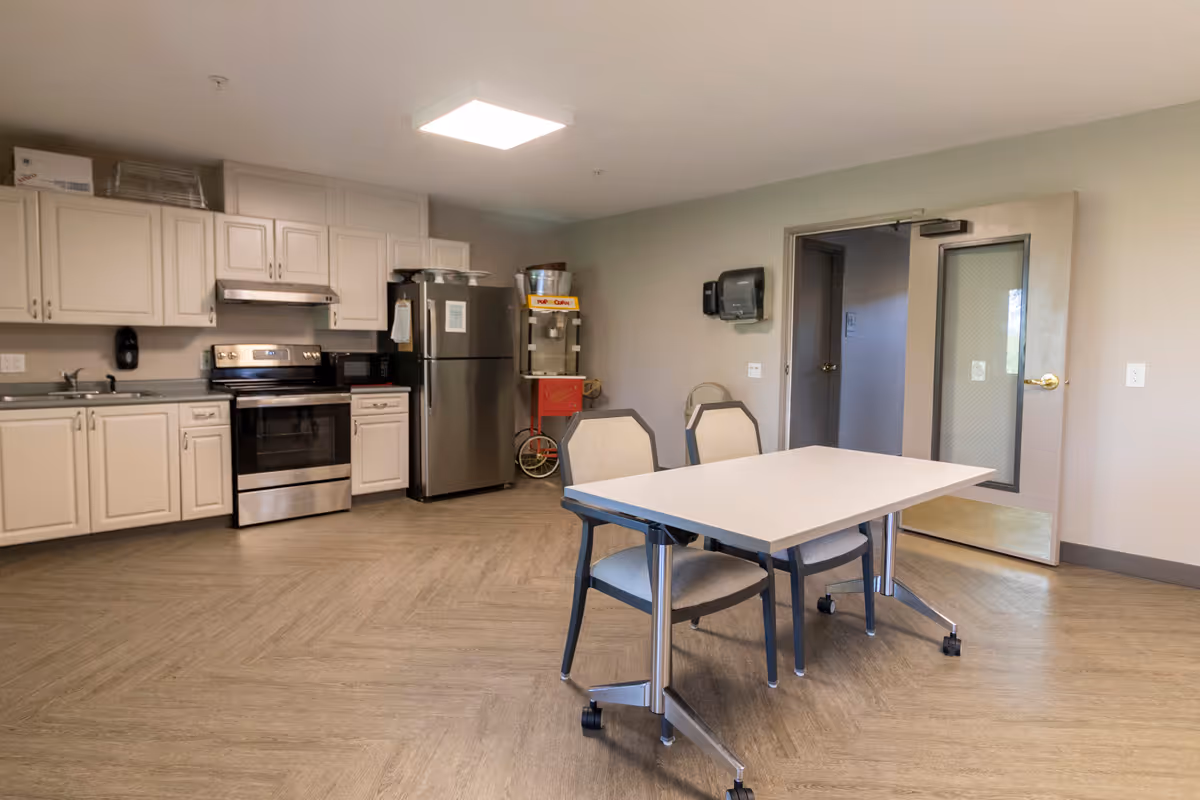 A clean kitchen area with white cabinets, a stainless steel stove and refrigerator, a microwave, and a popcorn machine. In the foreground, there is a white table with two chairs on wheels. The room has light-colored walls and flooring, with a door leading to another room.