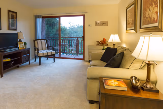 A cozy living room with a beige sofa adorned with black pillows, a wooden side table with a lamp and a book, a striped armchair, a wooden TV stand with a flat-screen TV, and a sliding glass door leading to a balcony with outdoor seating and greenery visible outside.