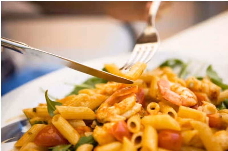 Close-up of a plate of pasta with shrimp, cherry tomatoes, and greens being cut with a fork and knife.