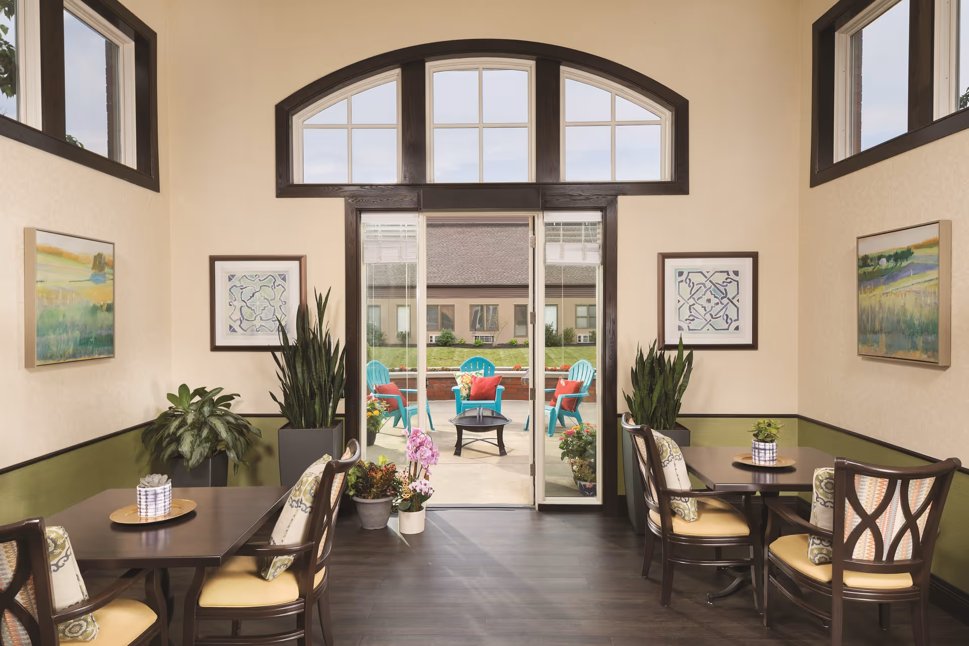 A bright and inviting interior seating area with two dark wooden tables, each surrounded by four cushioned chairs. The walls are decorated with framed artwork and potted plants are placed around the room. Large windows and a glass door lead to an outdoor patio with turquoise chairs and red cushions arranged around a fire pit.