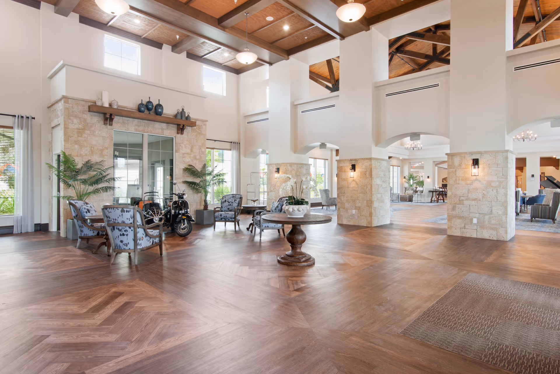 Spacious and well-lit senior living facility common area with high ceilings featuring wooden beams, large windows, stone pillars, and wood-patterned flooring. The room is furnished with patterned armchairs arranged in small seating groups, a round wooden table with a floral centerpiece, and decorative plants. The area has a warm and inviting atmosphere with soft lighting fixtures and visible adjoining rooms.