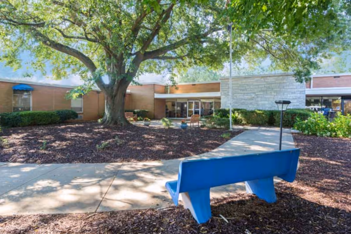 Outdoor view of Glenwood Health Center showing a large tree with green leaves, a blue bench, a concrete walkway, and the building entrance in the background surrounded by bushes and plants.
