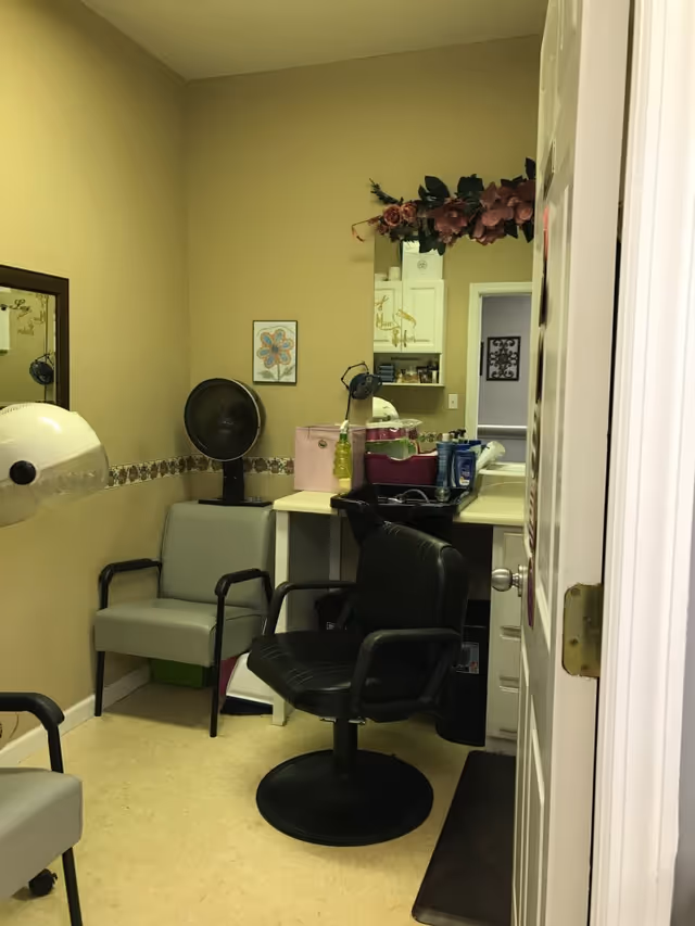 Interior view of a small hair salon or beauty station with a black salon chair, a gray waiting chair, a hair dryer hood, a counter with various hair products, and a mirror decorated with a floral garland.