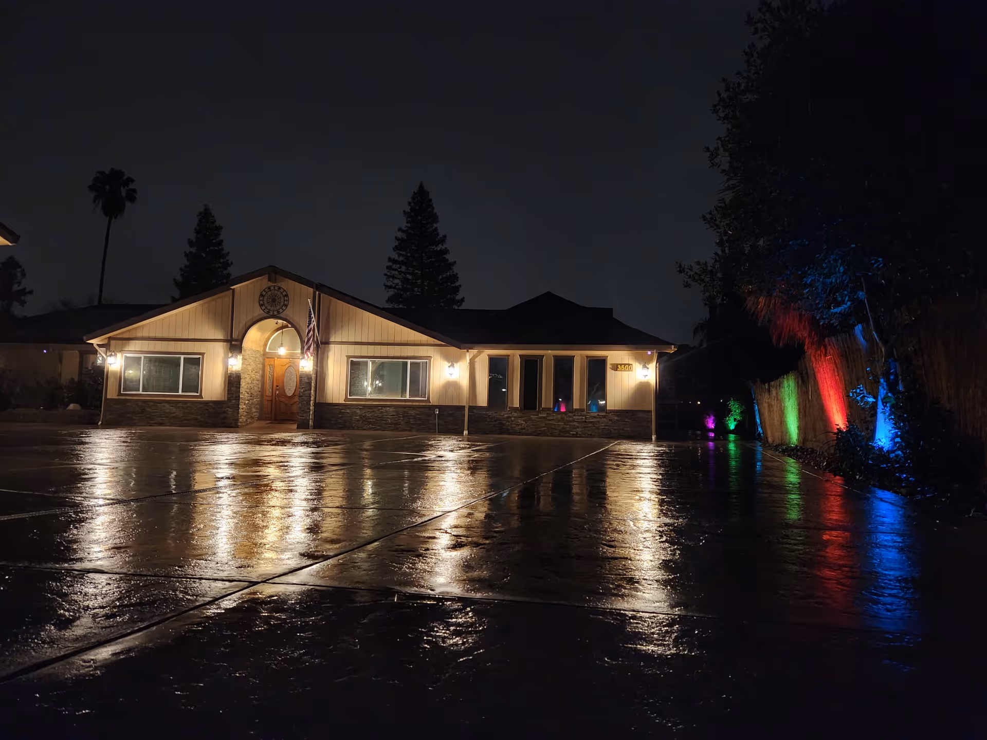 Nighttime view of a single-story building with exterior lights illuminating the entrance and windows. The wet pavement in front reflects the lights, and colorful lights in red, green, blue, and purple illuminate the trees and bushes along the right side of the building.