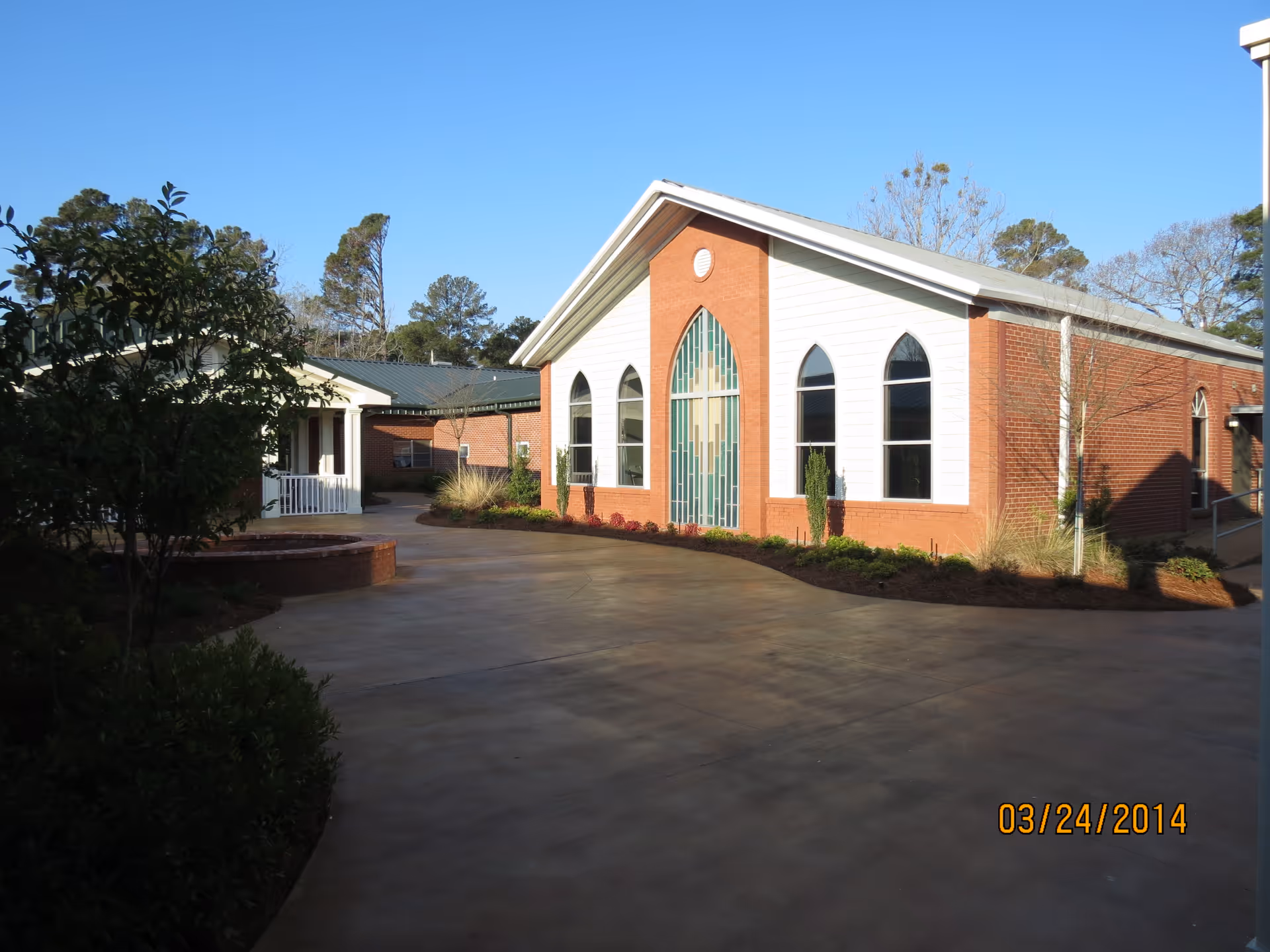 Exterior view of a brick building with white siding and arched windows, surrounded by a paved walkway and landscaped greenery under a clear blue sky.