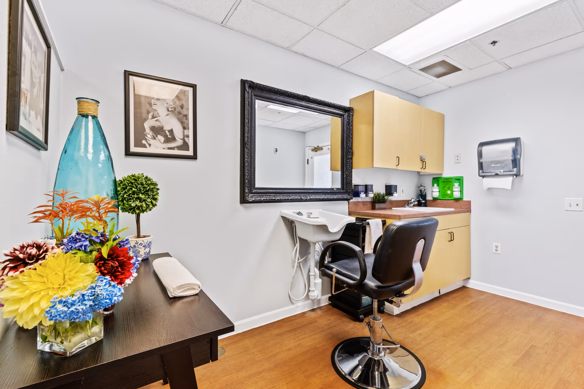A small salon or grooming room with a black salon chair in front of a white sink and a large black-framed mirror on the wall. There are light wood cabinets above a countertop with various items, including a green box and a paper towel dispenser mounted on the wall. A dark wooden table holds a colorful bouquet of flowers, a small potted plant, and a large blue decorative bottle. Two black and white framed photos hang on the wall.