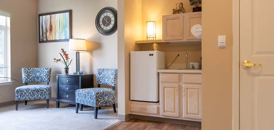 A cozy interior space featuring two patterned blue chairs with a small black side table between them, topped with a lamp and a flower vase. On the wall above the table is a colorful abstract painting and a round clock. To the right, there is a small kitchenette area with light wood cabinets, a white mini refrigerator, a sink, and a decorative lamp on the countertop.