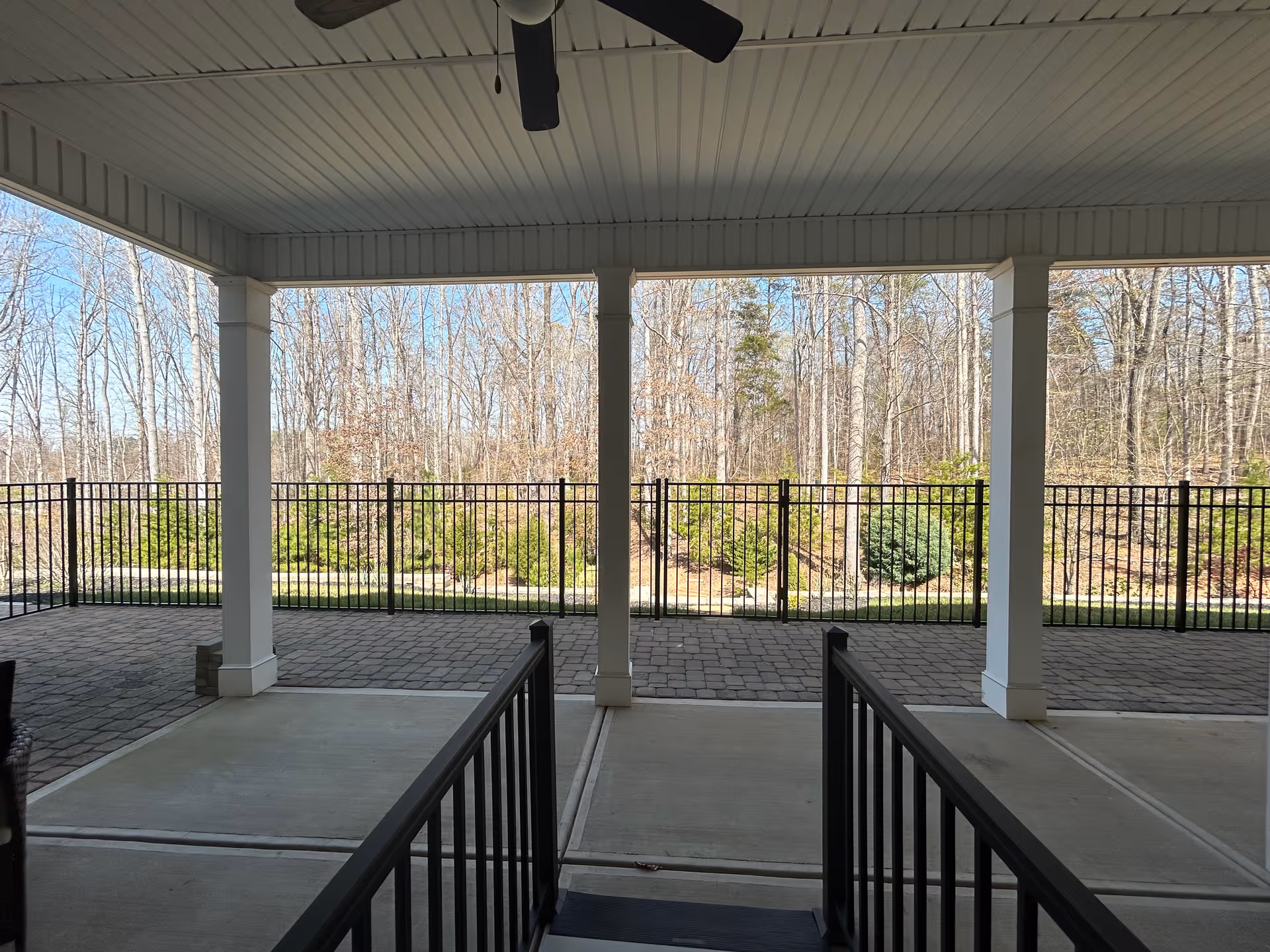 Covered outdoor patio area with a ceiling fan, white pillars, and black metal railings. Beyond the patio is a paved walkway and a black metal fence enclosing a landscaped area with bushes and leafless trees in the background under a clear blue sky.