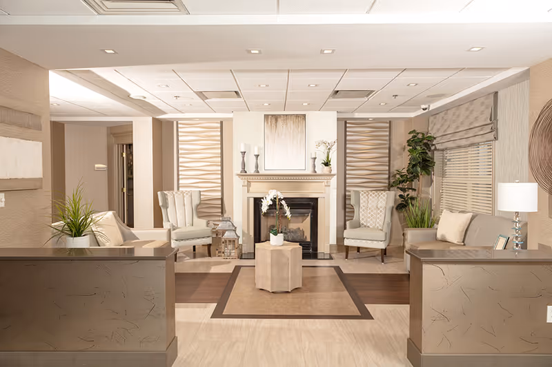 A cozy assisted living facility common area with a central fireplace flanked by two armchairs. The room features neutral tones with beige walls, a small table with a white orchid in front of the fireplace, decorative candles on the mantel, and plants on either side. There are two reception desks on the left and right sides of the image, and a window with blinds and a lamp on the right.