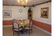Small dining room with a round table set for four under a chandelier, framed artwork, a wall clock, and a corner shelving unit with a plant.