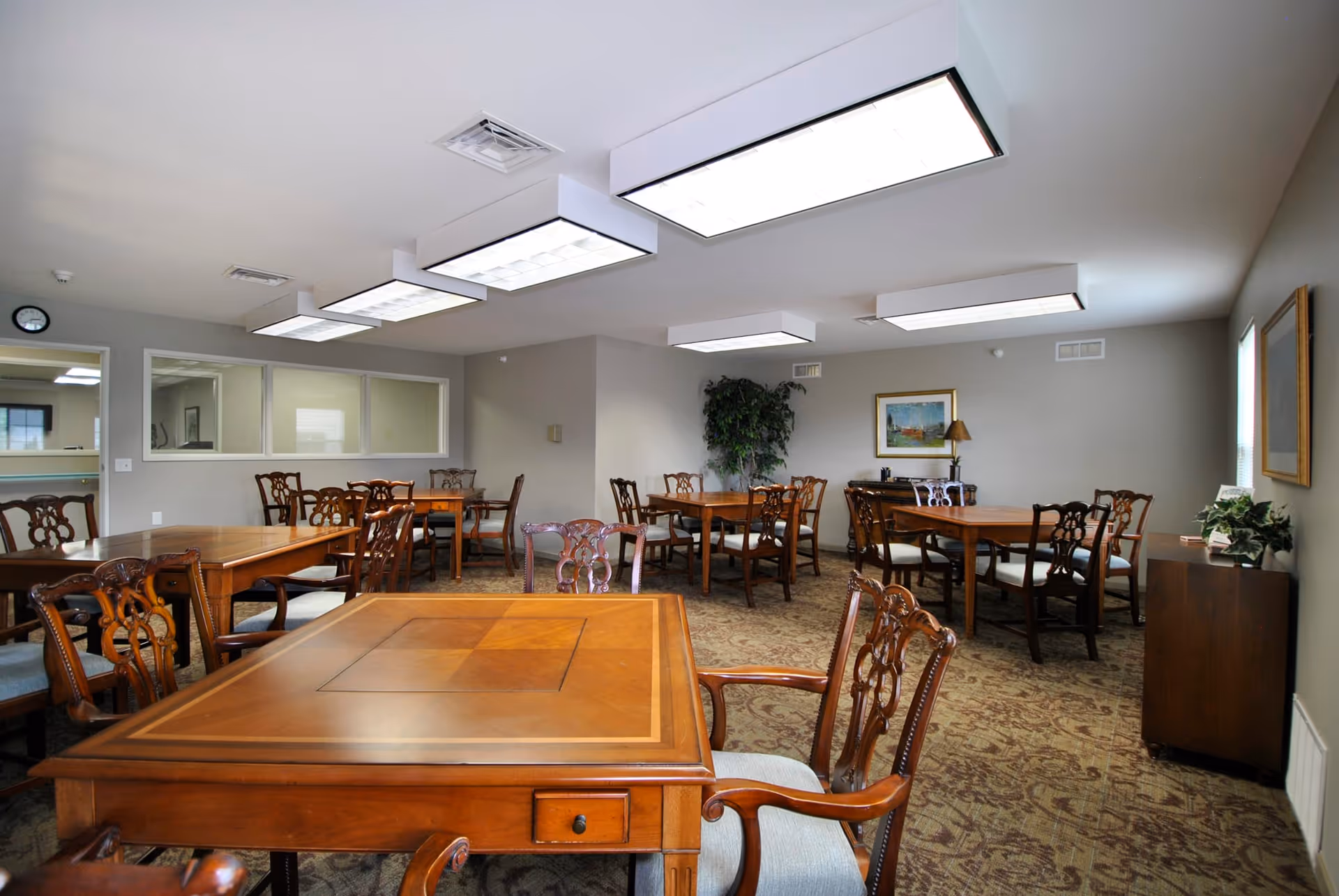 A well-lit dining room with multiple wooden tables and chairs arranged neatly. The room has beige walls, a patterned carpet, and several large rectangular ceiling lights. There are framed pictures on the walls and a potted plant in the corner.
