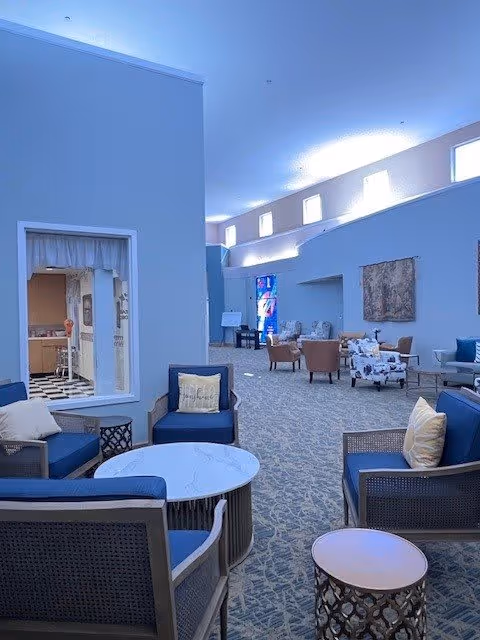 A spacious senior living community common area with blue cushioned chairs and small round tables arranged on a patterned carpet. The room has high ceilings with small windows near the top, and a kitchen area is visible through a window opening on the left side. Additional seating with armchairs and a sofa is seen further back in the room.