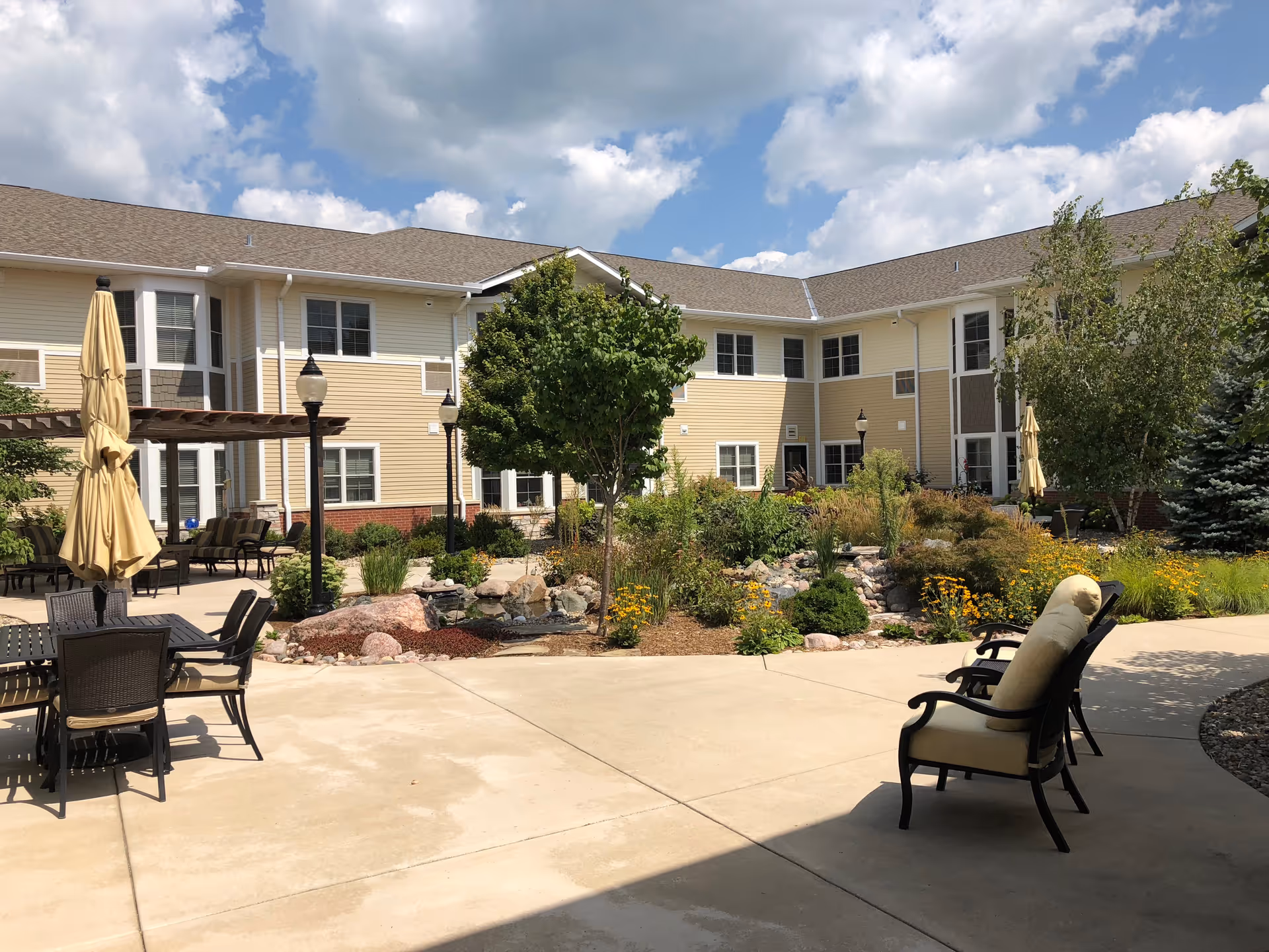 Outdoor courtyard area of a senior living facility with patio chairs, tables with umbrellas, a pergola, landscaped garden with rocks, flowers, and trees, and a two-story beige building in the background under a partly cloudy sky.