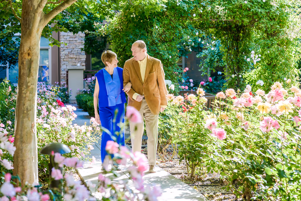 An elderly couple walking arm in arm along a garden path surrounded by blooming pink and white flowers and lush greenery, with a building visible in the background.